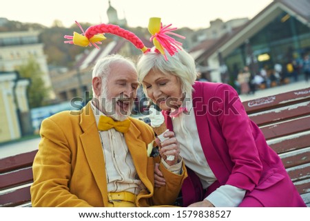 Jolly aged man and woman sitting together on bench in city and eating one dessert for two