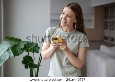 Jolly cheery friendly smiling positive middle-aged European woman standing near window, holding mobile phone, messaging, typing, looking into street, waiting for guests, online shop delivery courier