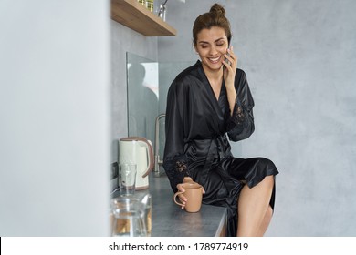 Jolly Female In Black Dressing Gown Is Sitting On Table With Cup Of Hot Drink And Talking On Smartphone