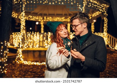 A jolly romantic couple is standing on city street and hugging on christmas eve while looking at each other. Portrait of a festive couple holding sparklers in hands and celebrating new year's eve. - Powered by Shutterstock