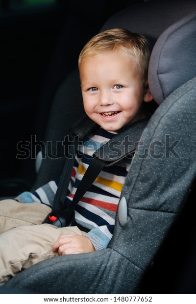 Jolly smiling preschool\
little blond boy is happily sitting in a safety chair, ready to\
ride in a family car. He\'s exited about his experience. Looking at\
camera.