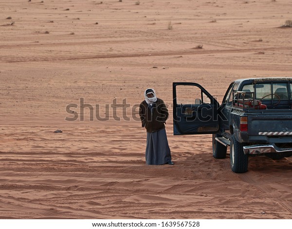 Jordan, Wadi Rum village - January 06, 2020: Old\
bedouin guide-driver stands near pickup truck with open door and\
waits for tourists in Wadi Rum desert in Jordan. Theme of travel\
and safari in Jordan.