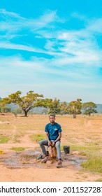 Jos East, Plateau State - May 12, 2021: Young African NGO Representative Visiting Rural Communities For Clean Water Projects On A Sunny, Bright And Blue Landscape Standing By A Well