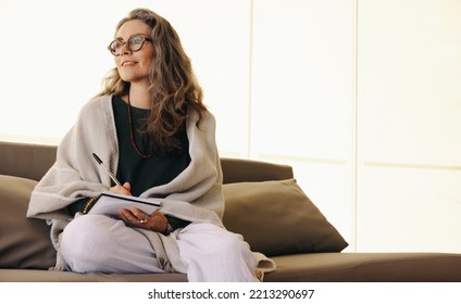 Journaling and self-awareness. Mature woman looking away thoughtfully while writing in her journal at home. Senior woman writing down her thoughts while sitting on a couch.