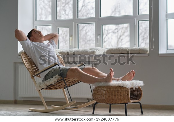 Joy of life. Total relaxation. Handsome\
young man keeping eyes closed and holding hands behind head while\
sitting in big comfortable chair at\
home
