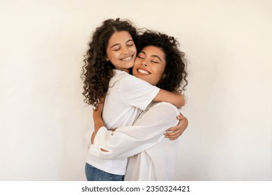 Joy of motherhood. Happy latin mother and daughter embracing, bonding and smiling over light wall background. Loving mom hugging her kid - Powered by Shutterstock