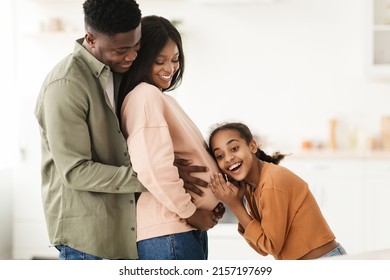 Joy Of Pregnancy. Happy Black Family Hugging Expecting Baby, Daughter Listening To Pregnant Mommy's Belly Standing In Modern Kitchen At Home. Childbirth And Parenting Concept