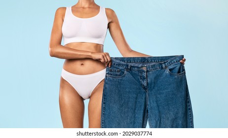 Joy Of Weight Loss. Closeup Of Fit Lady In White Underwear Holding Oversized Jeans Too Big After Great Weightloss And Successful Slimming Standing Posing At Blue Studio. Dieting Motivation Concept