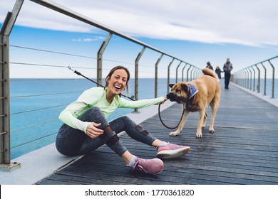 Joyful 40 Years Old Female In Sport Clothes Playing With Leash And Dog And Laughing While Looking At Camera On Pier