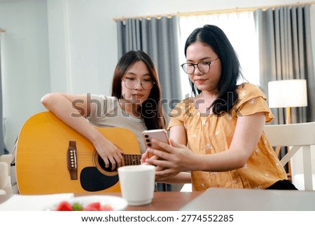 Joyful activity creating content at home by two Asian girls singing playing guitar : female friend sits and plays her favorite song classical guitar while friend takes pictures and the other girl happily watches the recorded performance.