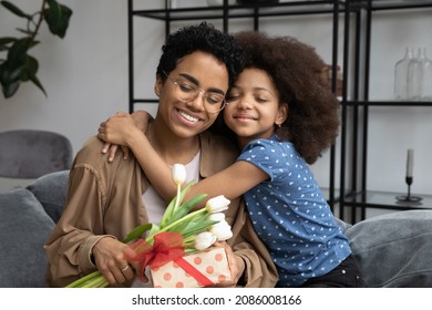 Joyful adorable small African American child girl cuddling loving young mother or nanny in eyewear, congratulating with happy birthday or International Women s Day, giving wrapped gist and flowers.