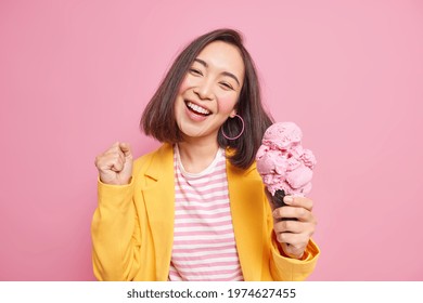 Joyful adorable teenage girl with eastern appearance dark hair tilts head clenches fist eats tasty ice cream smiles broadly has fun enjoys summer dressed in stylish clothes isolated on pink wall - Powered by Shutterstock