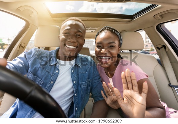 Joyful african american couple taking selfie while\
having car trip, sitting in auto, hugging and cheerfully smiling at\
camera, pretty millennial black lady embracing boyfriend and\
waving, sun flare