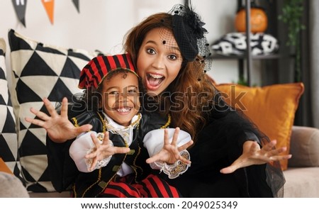 Joyful african american family mother and little boy son in Halloween costumes making scary gesture and looking at camera while sitting on sofa in decorated living room, celebrating all hallows day
