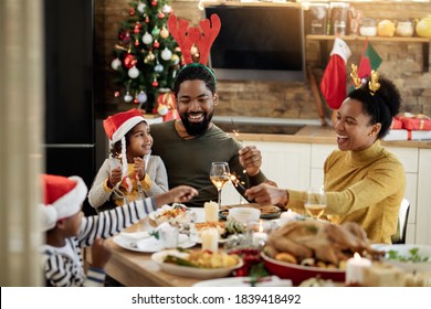Joyful African American family using sparklers while having lunch and celebrating Christmas at dining table. 