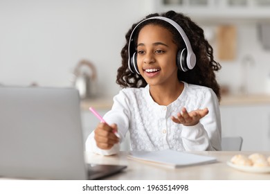 Joyful african american girl having online lesson from home, using wireless headphones and laptop, kitchen interior. Cute black schooler having video chat with teacher, copy space