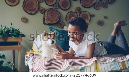 Joyful African American girl is using smartphone lying on bed and smiling stroking her cute dog with love, joy and tenderness. Modern technology, people and pets concept.