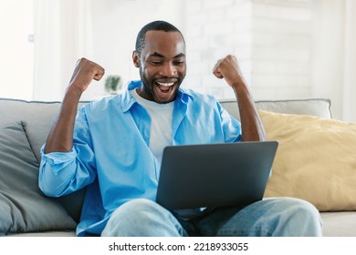 Joyful African American Guy Using Laptop, Working Online And Shaking Fists, Receiving Great News, Sitting On Sofa At Home. Distance Work And Freelance Career Concept