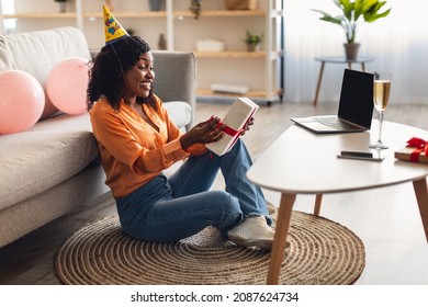 Joyful African American Lady Celebrating B-Day Online At Laptop Holding Wrapped Birthday Gift Sitting On Floor During Video Call With Friends At Home. Virtual Holiday Celebration. Side View