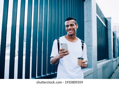 Joyful African American Male Looking At Screen Of Modern Cellphone While Standing Near Enclosure With Takeaway Hot Drink On Street