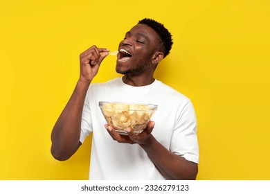 joyful african american man holding plate of chips and biting snack on yellow isolated background, the guy eats fast food from fried potatoes - Powered by Shutterstock