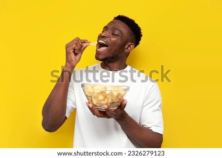 joyful african american man holding plate of chips and biting snack on yellow isolated background, the guy eats fast food from fried potatoes