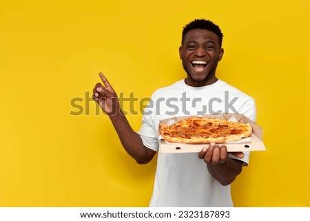 joyful african american man in white t-shirt holds box of pizza and points with his hand to the side on yellow isolated background, guy with fast food advertises copy space
