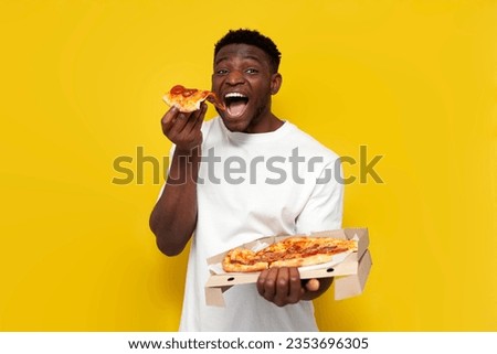joyful african american man in white t-shirt holds box of pizza on yellow isolated background, guy with fast food takes and eats slice of pizza