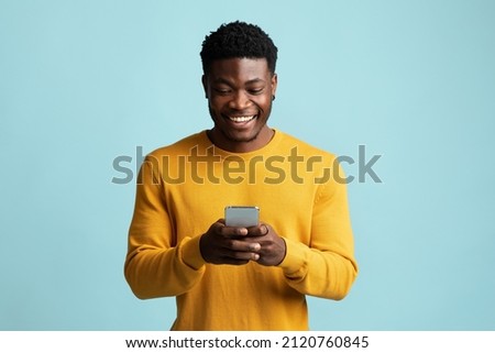 Joyful african american millennial guy holding modern smartphone, looking at gadget screen and smiling on blue studio background, black man gambling or gaming online, texting girlfriend, copy space