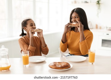 Joyful African American Mom And Daughter Eating Sandwiches Having Lunch And Drinking Orange Juice Sitting At Dining Table In Kitchen Indoors. Family Nutrition, Healthy Food Concept