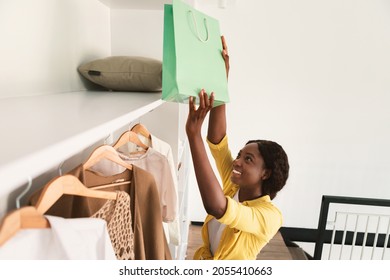 Joyful African American Shopaholic Lady Putting Shopping Bag In Wardrobe At Home. Fashion And Style, Shopaholism Concept. Female Taking Paper Shopper From Shelf In Closet. High Angle