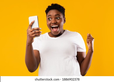 Joyful African American Woman Holding Smartphone Shaking Fists In Joy And Excitement Posing Standing Over Yellow Background. Yes, Great News Message Concept. Studio Shot