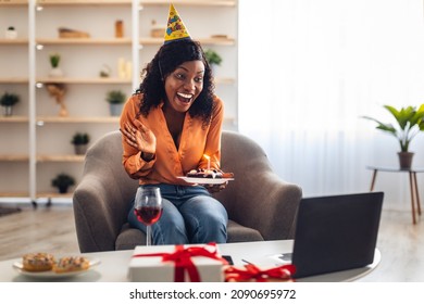 Joyful African American Woman Waving Hand To Laptop Celebrating Birthday Holding Plate With B-Day Cake And Wearing Festive Hat Sitting In Chair At Home. Online Party, Distance Celebration Concept