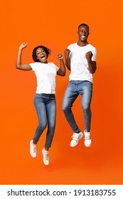 Joyful African American Young Couple Clenching Fists And Screaming, Jumping Up Over Orange Studio Background. Happy Black Millennial Man And Woman Celebrating Success, Full Length Photo