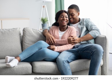 Joyful African American Young Couple In Casual Sitting On Couch, Hugging And Laughing, Living Room Interior, Copy Space. Happy Black Family Spending Cold Winter Day Together At Home, Cuddling