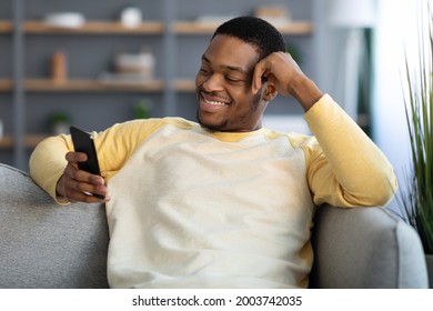 Joyful African American Young Man Chilling Alone At Home, Using Brand New Smartphone, Copy Space. Happy Black Guy Sitting On Couch In Living Room, Using New Dating App On Mobile Phone