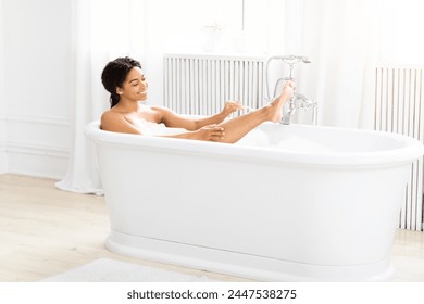 A joyful African American young woman enjoys a relaxing bubble bath in a white, freestanding bathtub in a bright, clean bathroom - Powered by Shutterstock