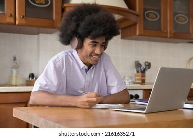 Joyful African high school student guy with natural curly hair wearing wireless headphones, writing study notes in copybook, watching learning webinar on laptop computer, making video call