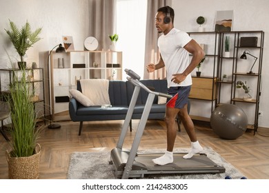 Joyful African Man In Modern Headphones Running On A Treadmill At Home. Male Runner Doing Cardio Exercise Healthily Lifestyle. Sporty Slender Man Sweating Workout In Apartment During Leisure Activity