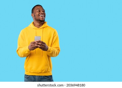 Joyful African Man Using Cellphone Texting And Looking Aside At Copy Space Posing Standing Over Blue Background, Studio Shot. Great New Mobile Application For Phone Concept - Shutterstock ID 2036948180