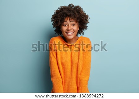 Joyful Afro American girlfriend gets unexpected surprise from boyfriend, has broad smile, feels pleased, wears orange jumper, expresses nice emotions, isolated over blue background. Facial expressions