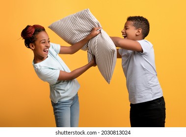 Joyful Afro American Siblings Brother And Sister Having Pillow Fight And Cheerfully Laughing, Yellow Studio Background. Happy Black Kids Having Fun Together, Enjoying Time Together. Childhood Concept