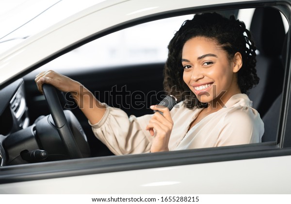 Joyful Afro Woman Showing Car Key Smiling To Camera\
Sitting In Driver\'s Seat In New Automobile Dealership Store.\
Selective Focus