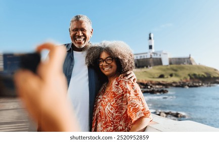 Joyful Afro-Brazilian couple poses for a selfie in front of the historic Farol da Barra lighthouse in Salvador, capturing memories on a beautiful coastal holiday. - Powered by Shutterstock