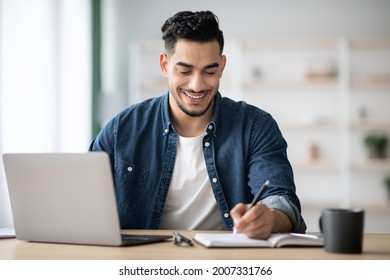 Joyful Arab Man Taking Notes While Working With Laptop At Office, Planning His Day In The Morning, Copy Space. Happy Middle-eastern Freelancer Looking For New Job Opportunities, Surfing On Internet