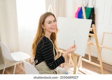 Joyful art instructor with paintbrush preparing to create artwork on a blank canvas while leading a painting tutorial in her studio - Powered by Shutterstock