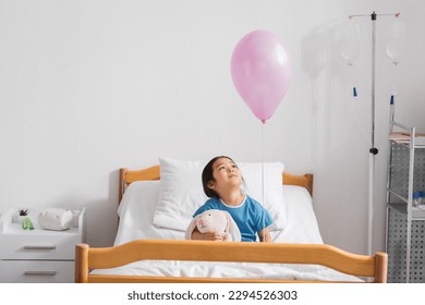 joyful asian girl sitting on hospital bed with toy bunny and looking at festive balloon - Powered by Shutterstock