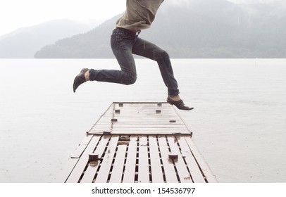 Joyful asian japanese young man jumping on a wooden pier while visiting a dreamy lake landscape with mountains during a cloudy and rainy day, outdoors.