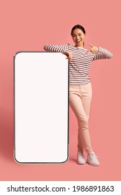 Joyful Asian Lady Leaning And Pointing At Big Smartphone With Blank White Screen, Demonstrating Copy Space For Your App Or Website Design, Standing On Pink Studio Background, Mockup Image