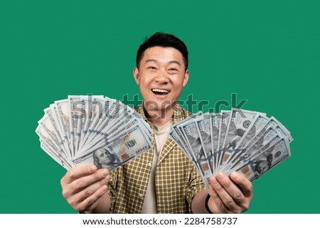 Joyful asian middle aged man holding bunches of money, celebrating success and lottery win over green studio background. Big luck, financial triumph, winning casino bet